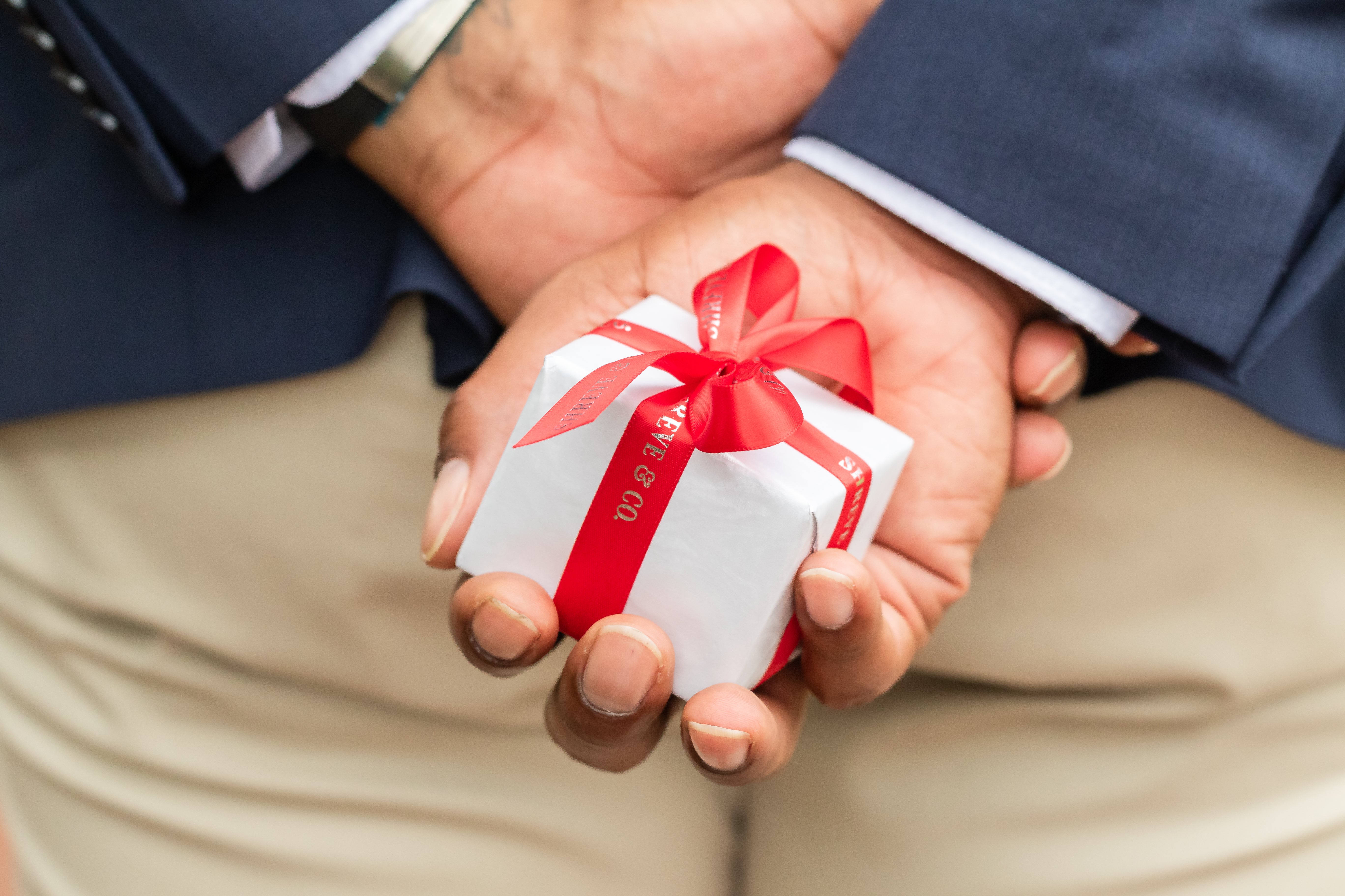 A man dressed elegantly, holding a ring box behind his back, symbolizing a romantic surprise or proposal. The setting conveys anticipation and a heartfelt moment.