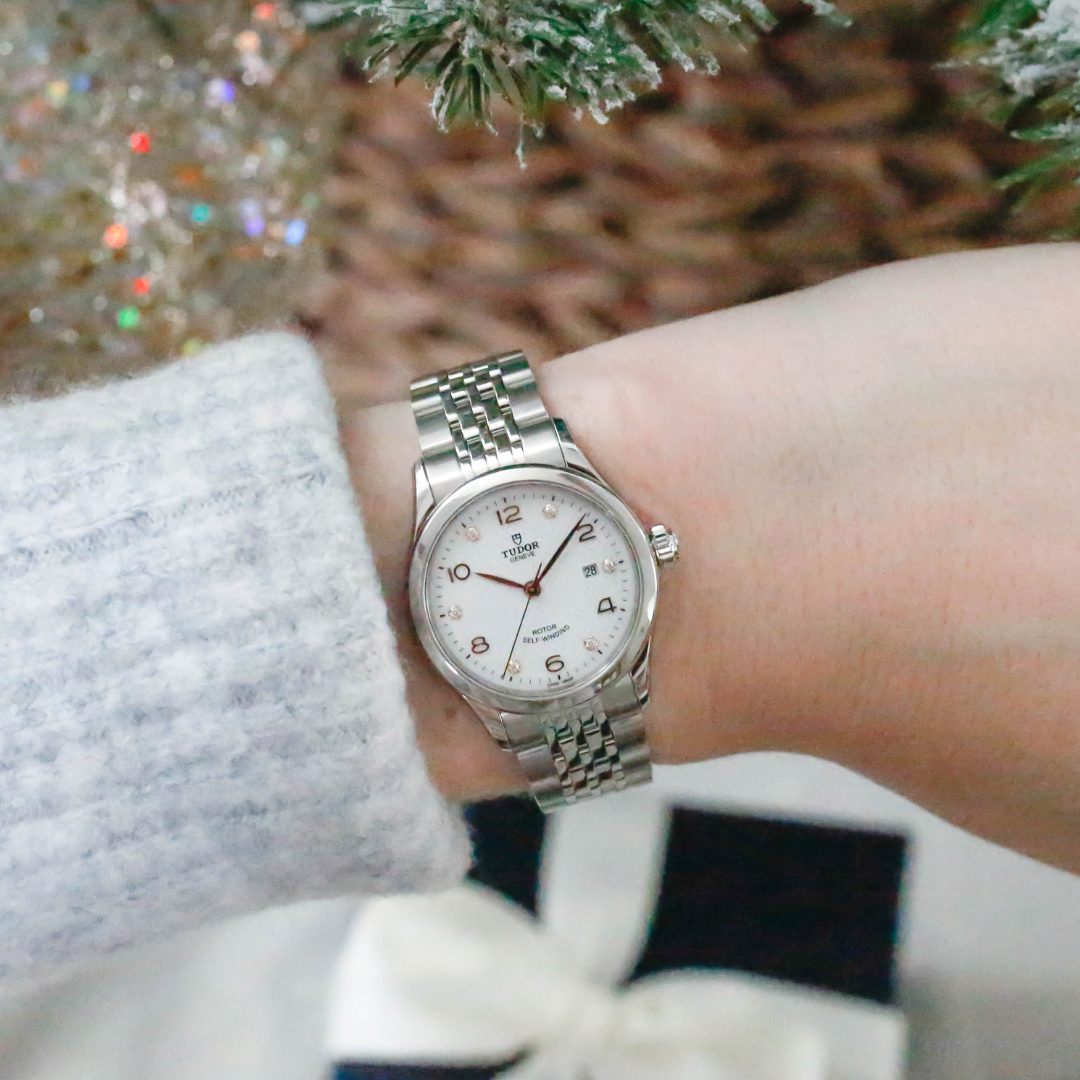 Close-up of a Tudor watch with a white dial and diamond markers on a wrist, set against a festive holiday background.